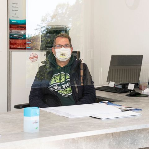 Fahrlehrer mit Mund-Nasen-Schutzmaske, hinter einer Spuckschutz Plexiglasscheibe mit Desinfektionsmitteln auf dem Schreibtisch